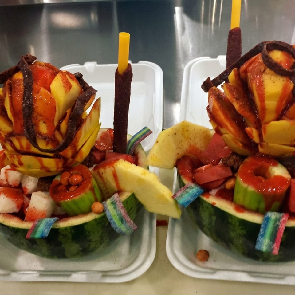 Two watermelon bowls filled with fruit, chili-coated candy, gummy strips, and carved fruit flowers on a kitchen counter.