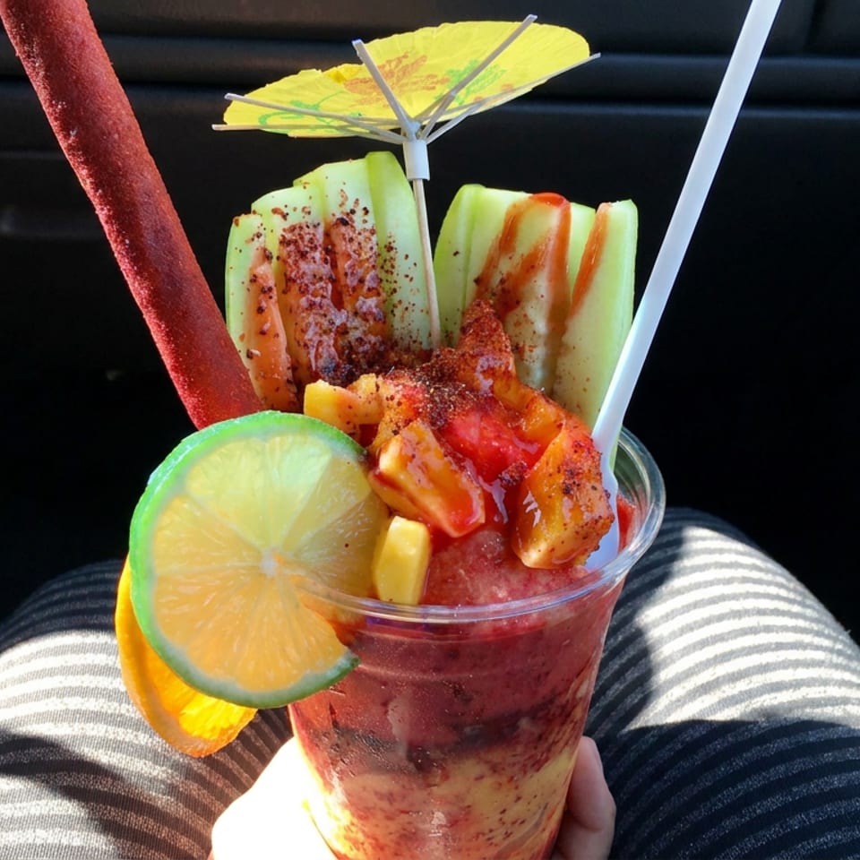 Spicy fruit cup with cucumber, lime, orange, chamoy, chili seasoning, and straws resting in a car.