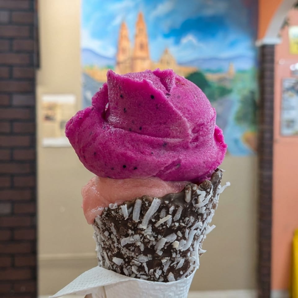 Colorful ice cream cone topped with vivid purple dragon fruit sorbet, chocolate coating, and shredded coconut inside a restaurant.