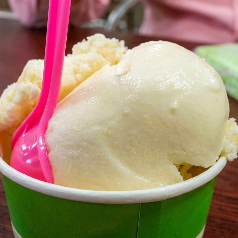 Scoop of pale green ice cream in a green cup with a bright pink spoon on a wooden table.