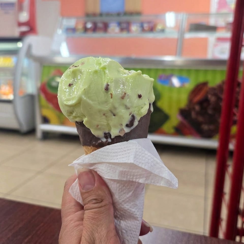 Hand holding a chocolate-dipped cone topped with a scoop of mint-green ice cream and chocolate pieces inside an ice cream shop.