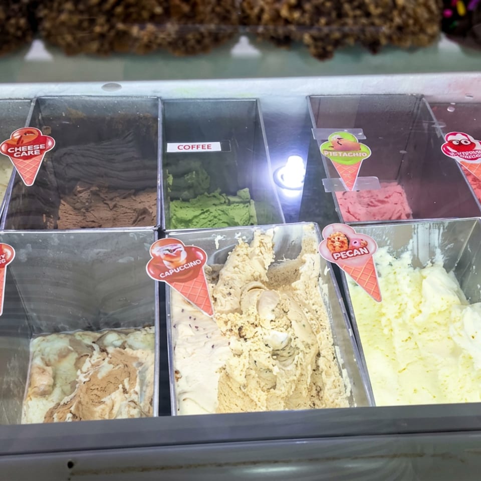 Display freezer filled with colorful gelato flavors, including pistachio, cappuccino, pecan, strawberry cheesecake, and chocolate.