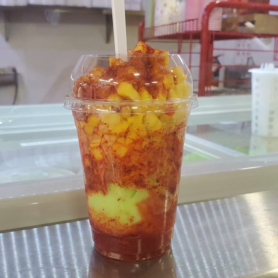 Clear cup filled with colorful fruit, chili seasoning, and a white straw on a restaurant counter.