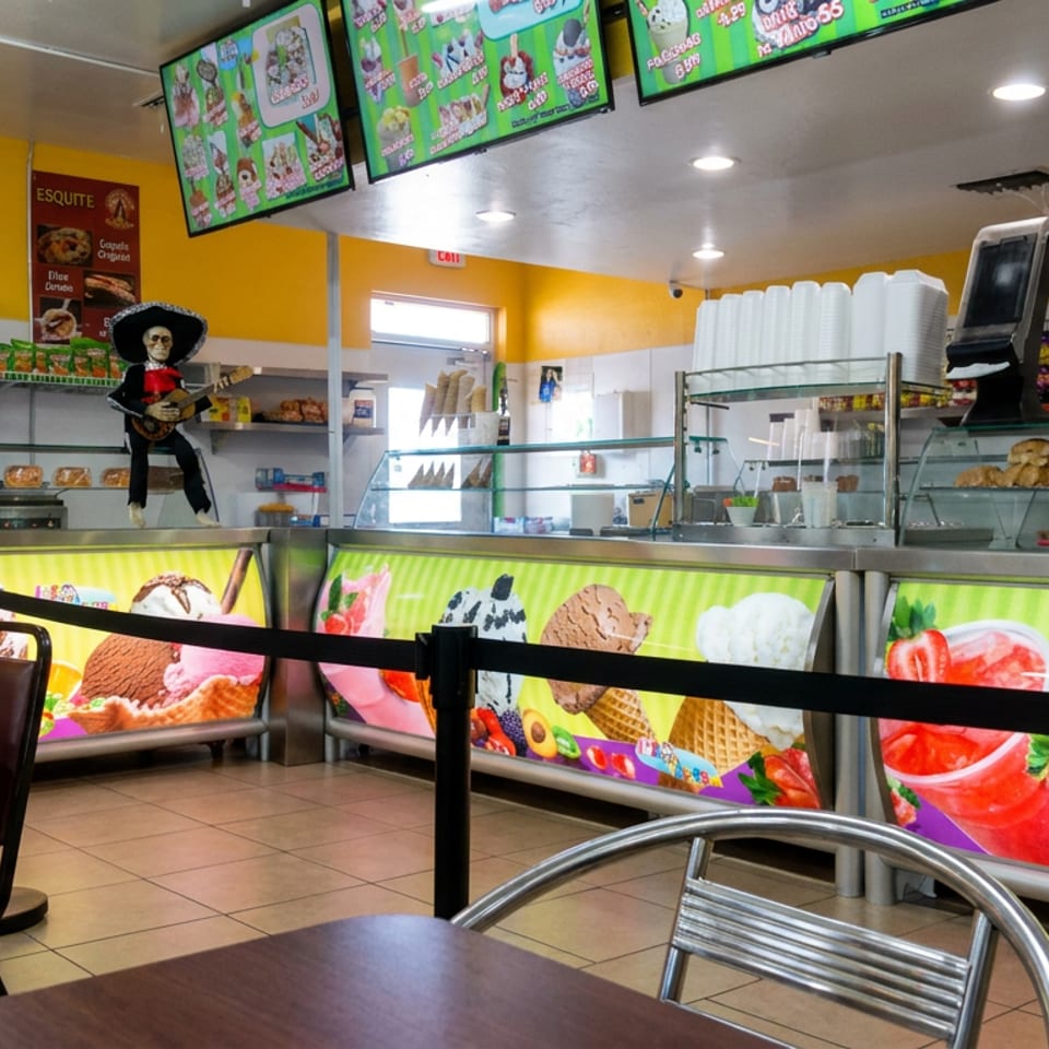 Bright ice cream shop interior with colorful dessert displays, digital menu screens, counter shelves, and café seating.