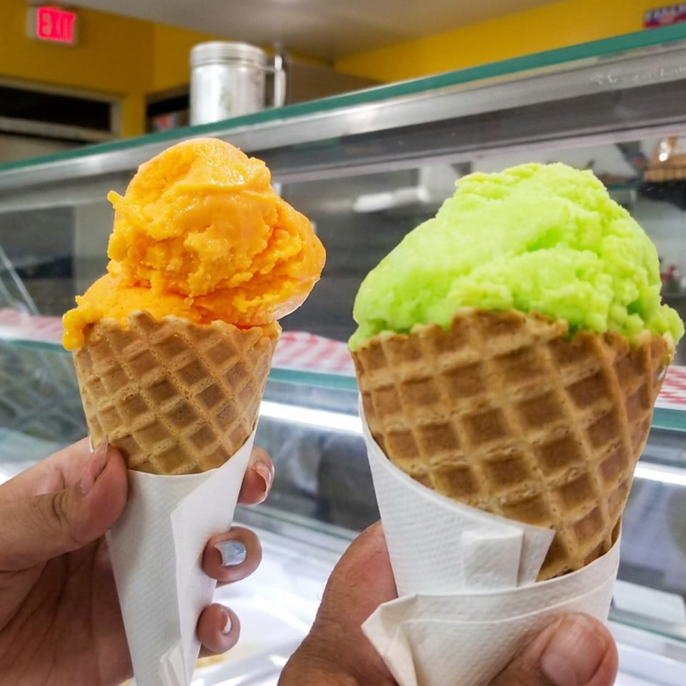 Two waffle cones with orange and green gelato held in front of a colorful ice cream shop display counter.