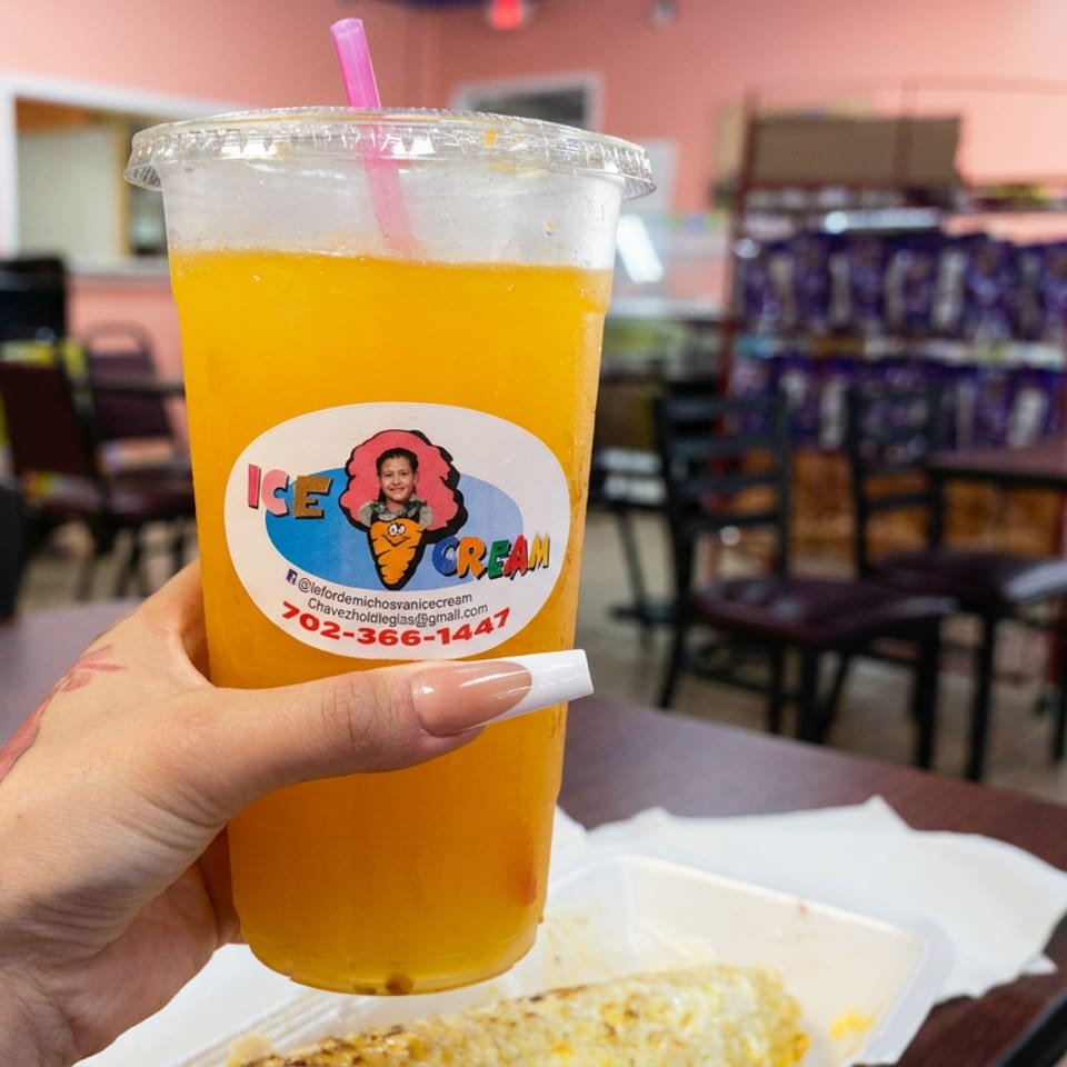 Hand holds a branded cup of bright orange frozen drink inside a casual ice cream shop, with tables and a beverage cooler in the background.